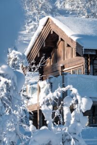 Sidan av timrat hus, balkongen syns bakom snötäckta träd